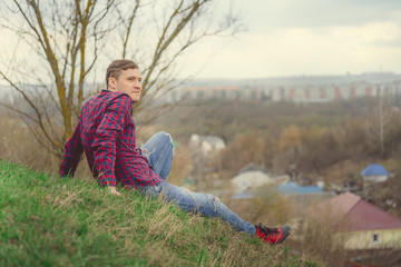 Naklejka na ściany i meble Young man sitting on green hill Side view of young male in jeans and plaid shirt sitting on grass and looking at camera