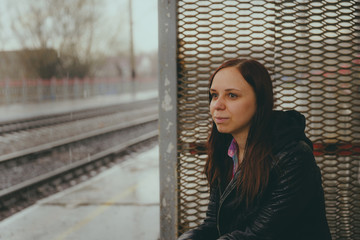 Dreamy young woman sitting beside railway Side view of young female in leather jacket sitting on background of railroad in overcast day
