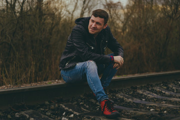 Trendy young man sitting on rails Pensive male in jeans and leather jacket sitting on railway in evening of cloudy day