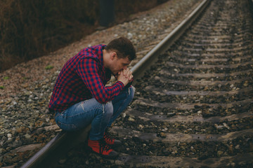 Trendy young man sitting on rails. Pensive male in jeans and red shirt on railway in evening of cloudy day