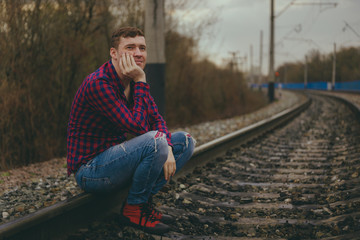 Trendy young man sitting on rails. Pensive male in jeans and red shirt on railway in evening of cloudy day