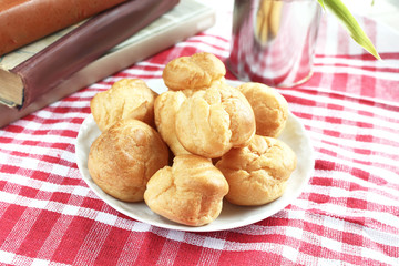 Homemade small cakes profiterole choux pastry with custard next to a white cup with tea and books on a table with a rustic tablecloth in a red-white cage