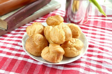 Homemade small cakes profiterole choux pastry with custard next to a white cup with tea and books on a table with a rustic tablecloth in a red-white cage