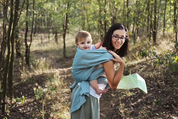European mother babywearing one year old on a hike in the forest, smiling and holding a map.