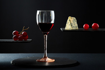 Composition of red wine in glass beside blue cheese and grape on black background.