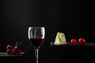 Composition of red wine in glass beside blue cheese and grape on black background.