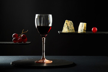 Composition of red wine in glass beside blue cheese and grape on black background.