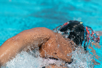 Fototapeta na wymiar Close-up of woman swimming freestyle. Sun tanned skin. Black swim cap, white tech suit. Blue pool water on sunny day. Splashing water.