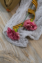 Two pink carnations, Saint George ribbon and military cap on a wooden surface.