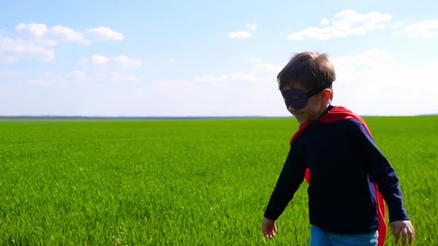 A happy joyful child in a superhero costume, a red cloak and a mask, runs across a green field and laughs