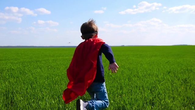A child dressed as a superman runs across a green field on a sunny day