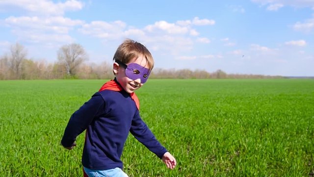 Joyful Child In A Superhero Costume, A Red Cloak And A Mask Runs Through The Green Grass, Smiling And Looking Toward The Camera