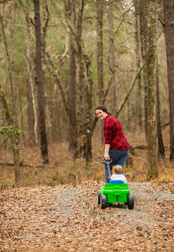 A Caucasian Mother Wearing A Black And Plaid Shirt Pulls Her One-Year Old Son In A Green Wagon On A Gravel Road Covered With Dead Leaves; New Hill, North Carolina, United States Of America