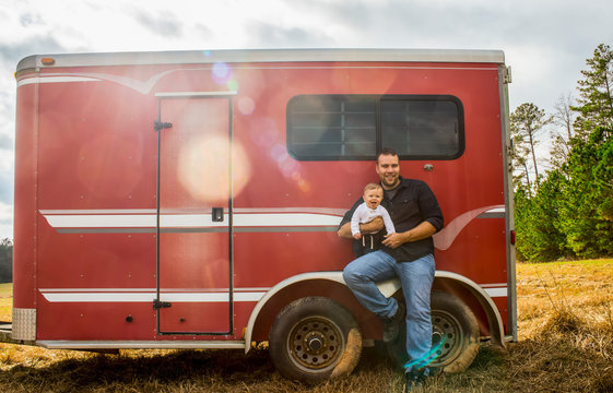A Baby Caucasian Boy And His Father Sit On The Wheel Well Of A Red Horse Trailer; New Hill, North Carolina, United States Of America
