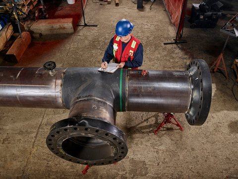A Tradesman Stands At A Large Pipe And Writes On A Document; Edmonton, Alberta, Canada