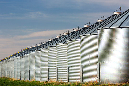 Close-Up Of A Long Row Of Shiny Metal Grain Bins Reflecting Sunlight With Blue Sky And Clouds; Beiseker, Alberta, Canada