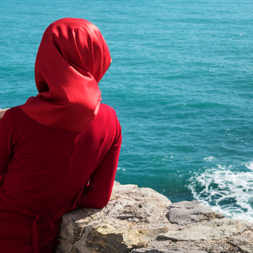 A woman wearing a red headscarf and red dress stands at a stone wall looking out at the blue water; Budva, Budva Municipality, Montenegro