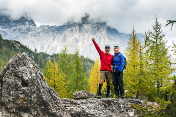 Male and female hikers standing on rock with autumn coloured trees and cloud-covered mountain range in the background; Sesto, Bolzano, Italy