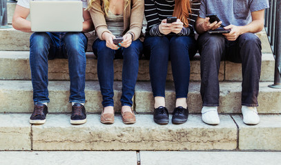 Four students sitting in a row on a step using their technology on the university campus, with a view of their feet and legs only; Edmonton, Alberta, Canada