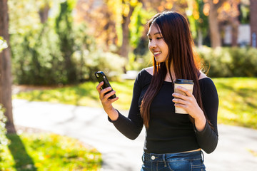 A young Chinese International student stands holding a coffee cup and using her smart phone on a university campus; Edmonton, Alberta, Canada