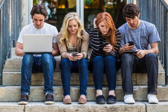 Four students sitting in a row on a step using their technology on the university campus; Edmonton, Alberta, Canada