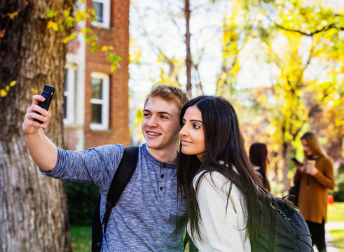 A couple who are university students posing for a self-portrait with a smart phone with a small group of friends in the background; Edmonton, Alberta, Canada