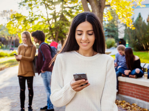 A young female university student of Lebanese ethnicity checks her smart phone while her friends are talking together in the background on the university campus; Edmonton, Alberta, Canada