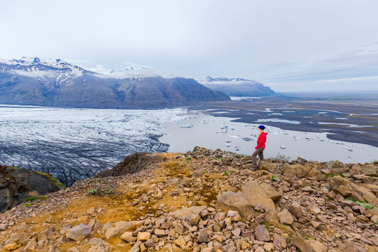 A female hiker in warm clothing for the elements stands on the edge of moutain cliff overlooking the glacier lake and valley views of Vatnajokull National Park; Iceland