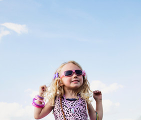 A young girl all dressed up with blond curly hair wearing sunglasses and jewelry standing making cute faces against a blue sky with cloud; Spruce Grove, Alberta, Canada