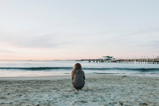 A girl crouches on the beach looking out over the ocean to the horizon at sunset; Long Beach, California, United States of America