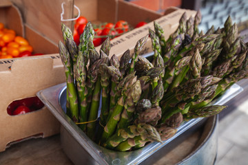 Lots of freshly picked asparagus tips sitting in a chefs metal gastro tin with fresh tomatoes in boxes behind