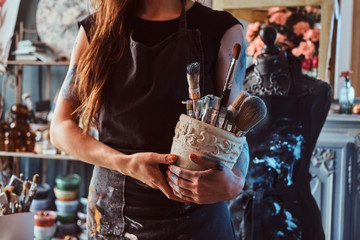 Young artist is tidying up after long working day in her own studio. She is holding jars with brushes.