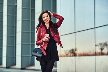 Beautiful smiling woman in red jacket is standing next to glass building with smartphone in her hands.