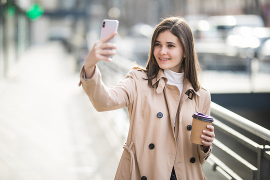 Beautiful young blonde woman wearing a coat walking outdoors, taking a selfie with mobile phone, holding takeaway coffee cup