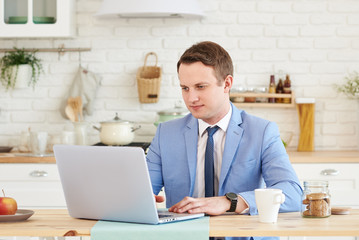 Handsome young businessman working on laptop computer at the table at home. drinking coffee at the kitchen in the morning.  Checks bills in accounting