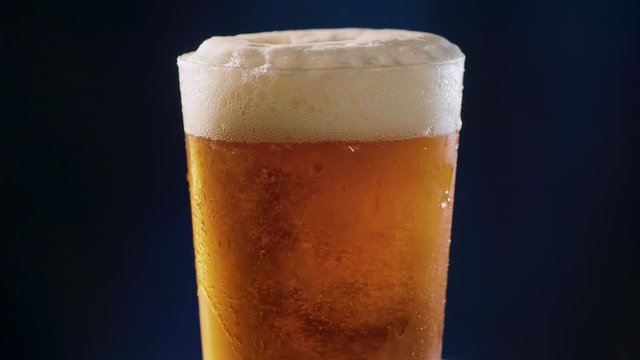 Golden Beer poured to a glass, rotating over a dark blue background, slow motion.