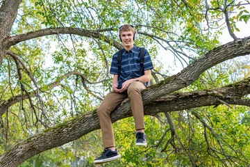 Happy student sitting on a tree branch while listening to music through his headphones.