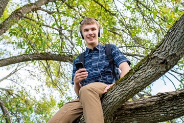 Happy student sitting on a tree branch while listening to music through his headphones.