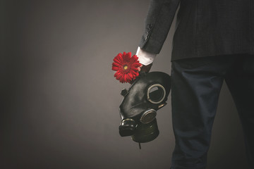 Man in gas mask and suit is holding in hands a red gerbera flower on a gray background. Pollution...