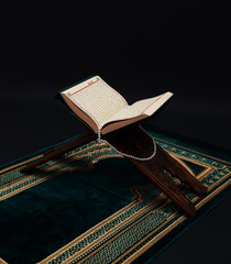 Islamic Holy Book Quran on wood carving rahle with rosary beads and prayer rug on black background....