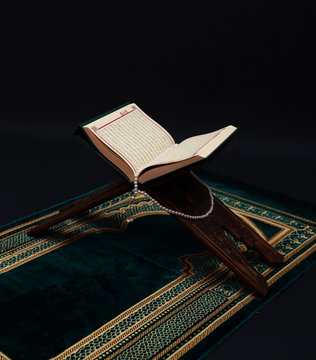 Islamic Holy Book Quran on wood carving rahle with rosary beads and prayer rug on black background. Kuran the holy book of Muslims. Ramadan concept.