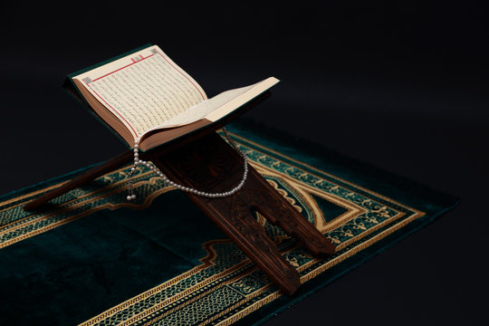 Islamic Holy Book Quran on wood carving rahle with rosary beads and prayer rug on black background. Kuran the holy book of Muslims. Ramadan concept.