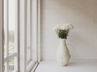 White chrysanthemums in ceramic jug next to window and against painted brick wall - vintage filter and selective focus