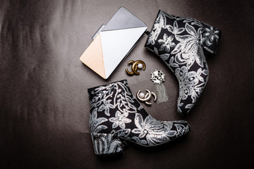 black ankle boots with thick heels decorated with flowers embroidered with silver sequins and a metal clutch and brooches on a gray paper background