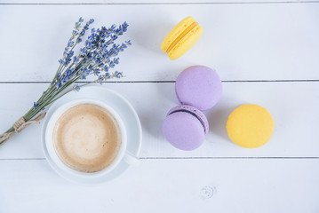 Fototapeta premium Sweet delicious violet and yellow macarons and cup of latte or americano and branch of fragrant lavender on white wooden background.