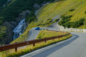 Light blue car races through a hairpin turn running past glassy mountain stream.