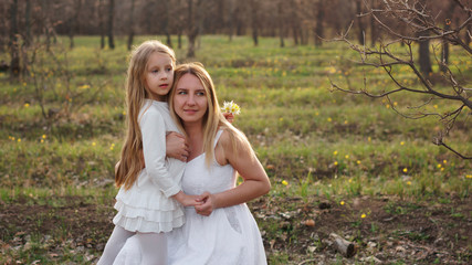 Fototapeta premium Family portrait of a single mother hugging her daughter. They are posing in white dresses against the backdrop of a spring meadow. Mothers Day. The girl has a bouquet of forest tulips in her hand
