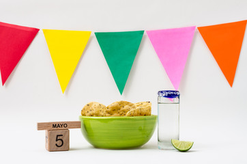 Wooden cube calendar with cinco de mayo date, green bowl with tortilla chips, tequila shot glass, lemon and colorful flags on white background