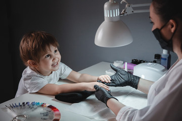 A little girl in a beauty salon. Looks at the manicurist in a black mask and black gloves. Nail...