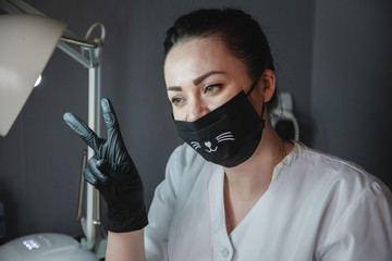 Manicurist in a black mask and black gloves sitting at work. Close-up portrait. The image of a...